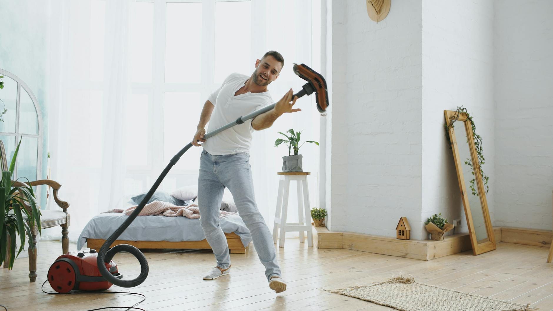 Joyful man using a vacuum cleaner as a microphone in a bright, tidy bedroom.