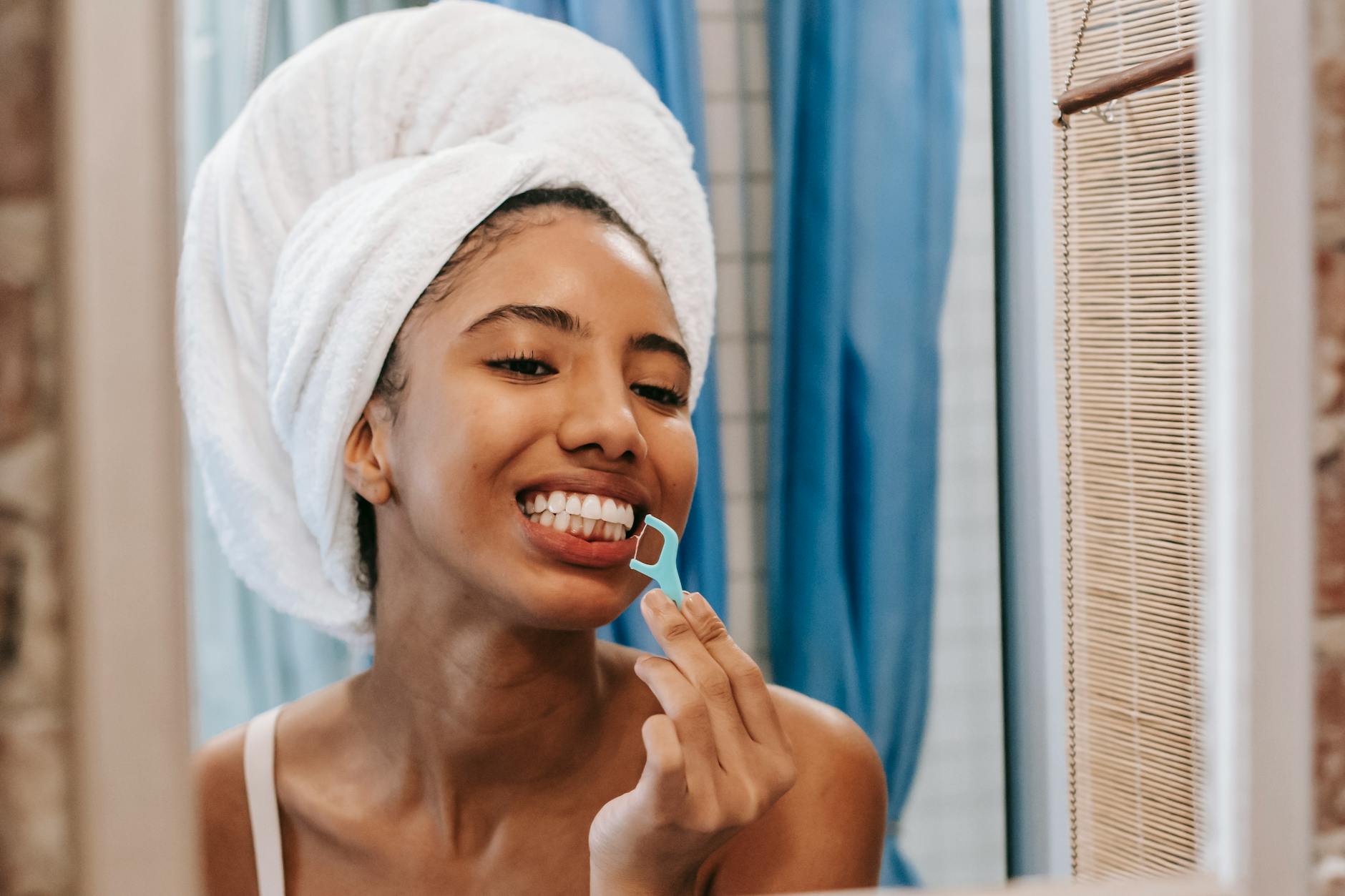 Young ethnic female with wet hair wrapped in towel turban cleaning teeth with flossier while looking at reflection in mirror