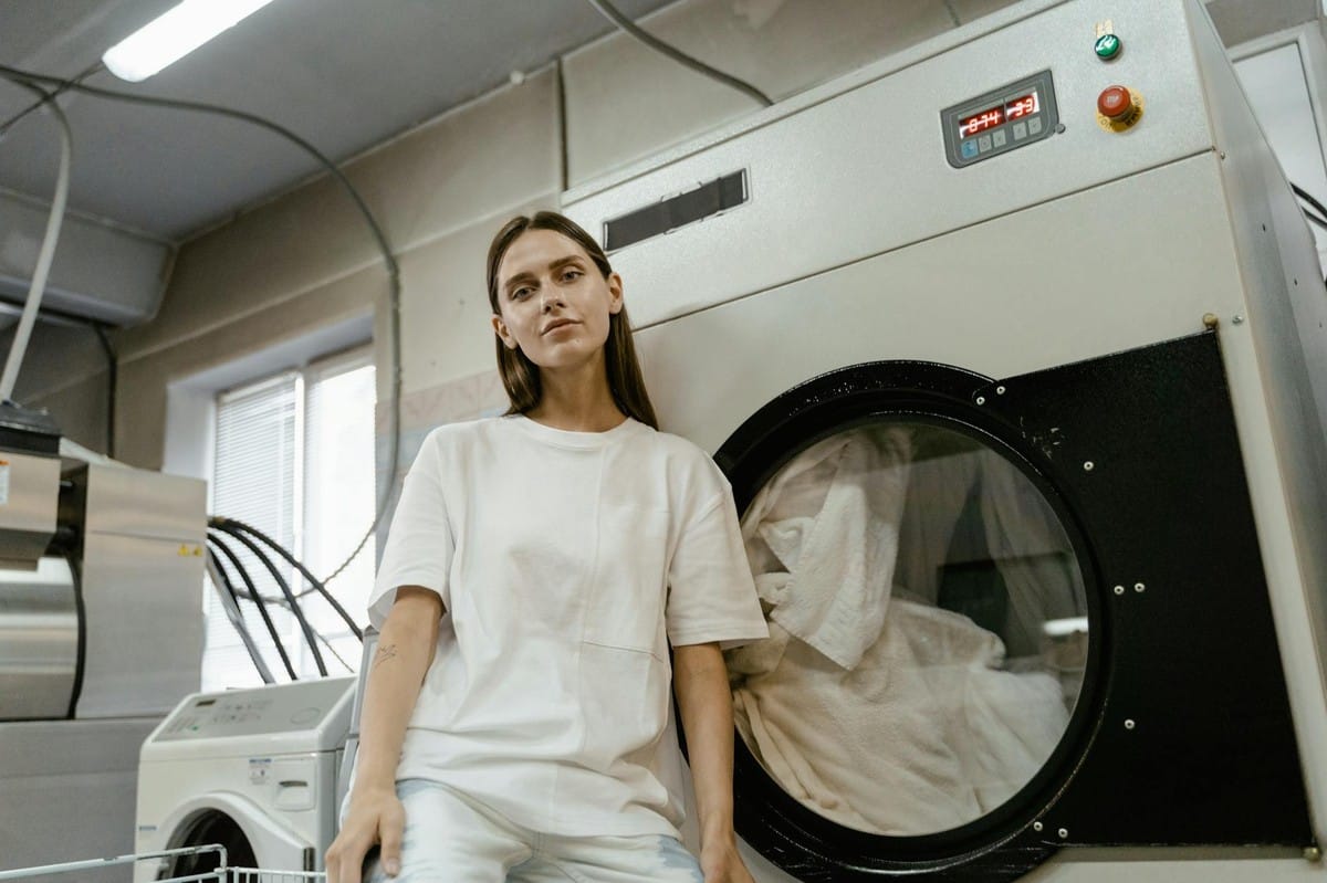 Young woman sitting in a laundry facility beside a large industrial washing machine.